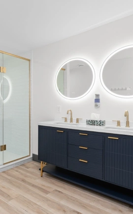 Beautiful bathroom featuring lit mirrors, a glass shower, and navy vanity with gold hardware.